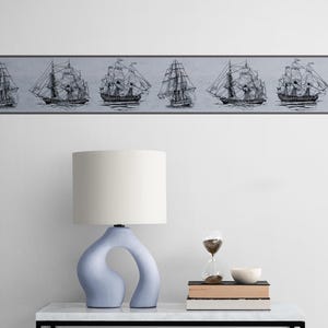 May include: A white lamp with a blue, abstract base sits on a white table with a stack of books and an hourglass. A border of black and white ship illustrations is on the wall behind the table.
