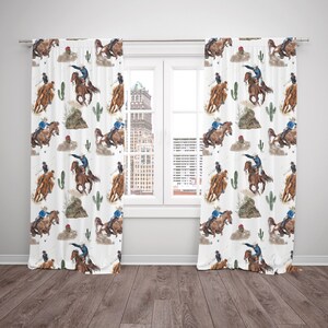 May include: White curtain panels with a repeating watercolor pattern of cowboys riding horses, cacti, and desert landscapes. The cowboys wear blue shirts and hats. The curtains are hanging in front of a window.