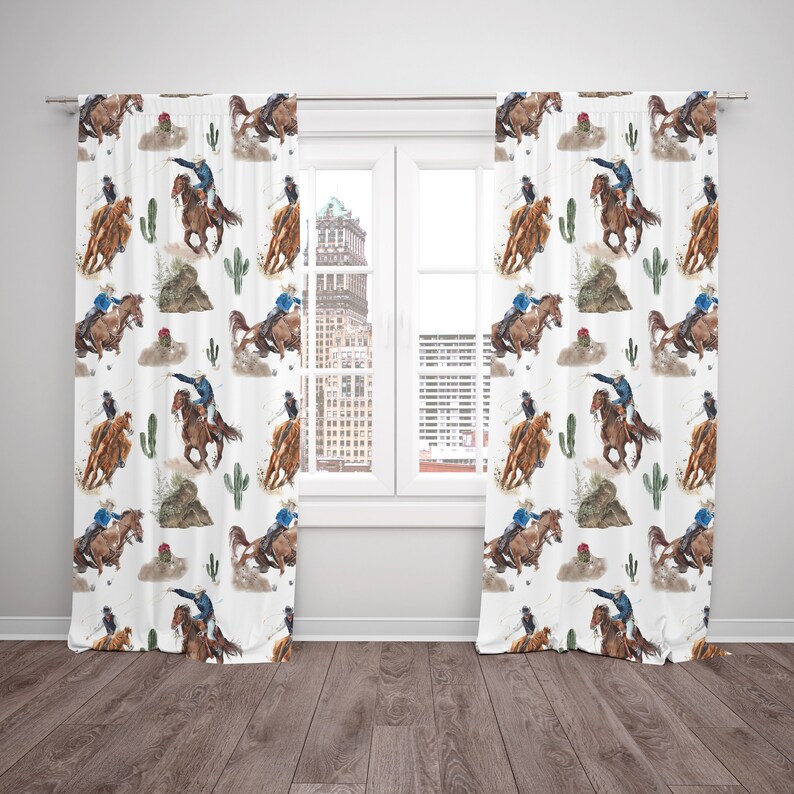 May include: White curtain panels with a repeating watercolor pattern of cowboys riding horses, cacti, and desert landscapes. The cowboys wear blue shirts and hats. The curtains are hanging in front of a window.