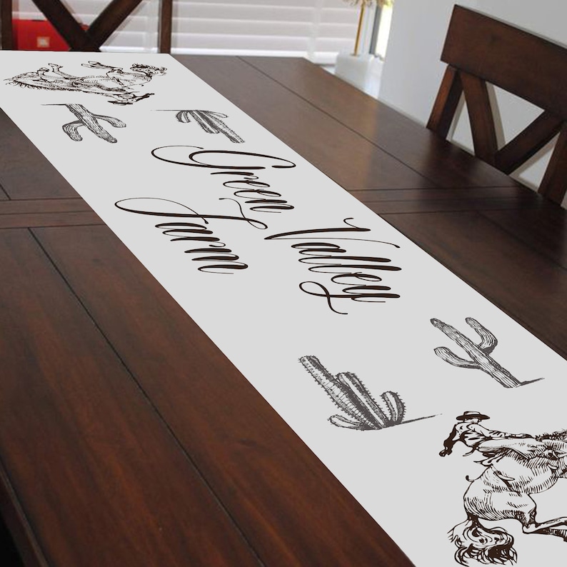 May include: White table runner with the text "Green Valley Farm" in brown cursive script. The runner is decorated with black and white illustrations of cacti and a cowboy riding a horse.