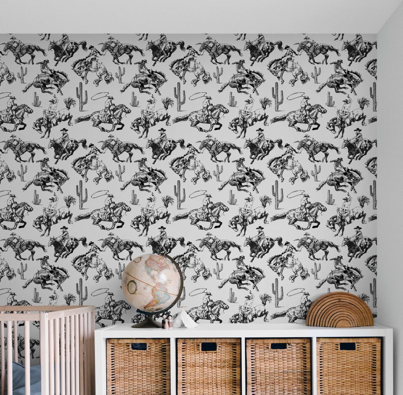 May include: Black and white wallpaper featuring a repeating pattern of cowboys riding horses. The cowboys are wearing hats and holding ropes. The horses are in various poses, some are jumping and some are running. The wallpaper is on a white wall in a room with a crib and a globe.