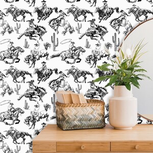 May include: Black and white wallpaper featuring a repeating pattern of cowboys riding horses. The cowboys are wearing hats and holding ropes. The horses are galloping and jumping. The background is white.
