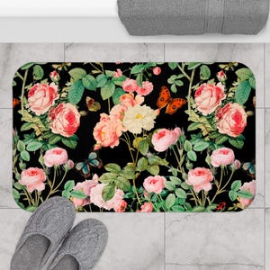 May include: A rectangular bath mat featuring a black background with a botanical print of pink and white roses, green leaves, and orange and blue butterflies. The mat has rounded corners and is positioned on a light gray tiled floor surface.