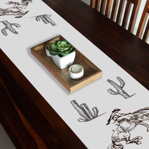 May include: White table runner with a brown cactus and cowboy riding a horse design. The runner is on a dark brown wooden table with a small potted plant and candles in a wooden tray.