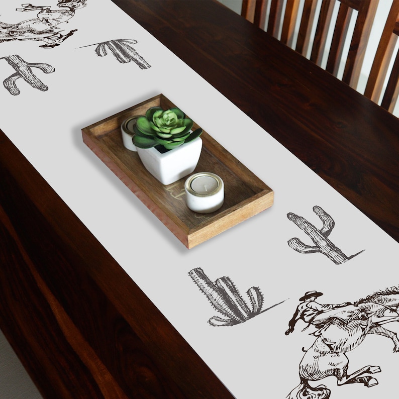 May include: White table runner with a brown cactus and cowboy riding a horse design. The runner is on a dark brown wooden table with a small potted plant and candles in a wooden tray.