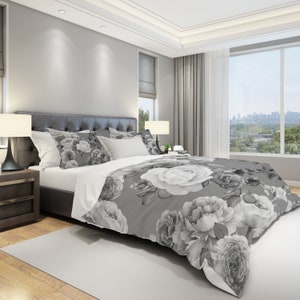 May include: A grey and white floral duvet cover with a large white rose in the centre. The duvet cover is on a bed with white sheets and grey pillows.