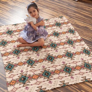 K&ouml;nnte beinhalten: Ein M&auml;dchen sitzt auf einer beige-braunen, gemusterten Wolldecke mit einem wei&szlig;en Pl&uuml;sch-Einhorn. Die Decke hat ein geometrisches Muster mit t&uuml;rkisfarbenen, orangen und rosafarbenen Akzenten.