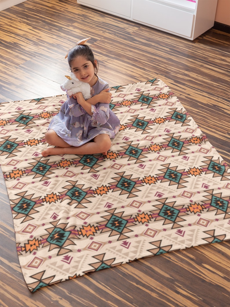 K&ouml;nnte beinhalten: Ein M&auml;dchen sitzt auf einer beige-braunen, gemusterten Wolldecke mit einem wei&szlig;en Pl&uuml;sch-Einhorn. Die Decke hat ein geometrisches Muster mit t&uuml;rkisfarbenen, orangen und rosafarbenen Akzenten.