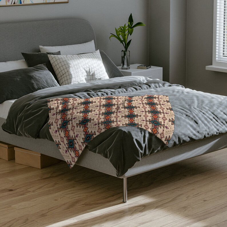 K&ouml;nnte beinhalten: Eine beigefarbene Tagesdecke mit einem geometrischen Muster in Orange-, Rot-, Blau- und Schwarzt&ouml;nen. Die Decke liegt auf einem grauen Bett mit Kissen.
