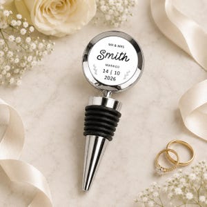 May include: A silver-toned metal bottle stopper with a circular top featuring the printed text MR & MRS Smith MARRIED 14 | 10 2026 and small leaf illustrations. The stopper has a black ribbed rubber base for sealing bottles and a tapered metal tip.