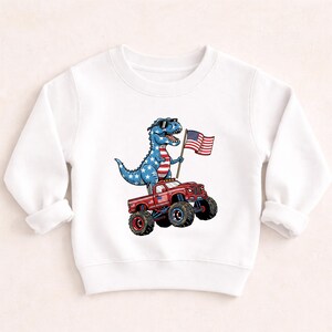 A white long-sleeved shirt with a graphic design depicting a dinosaur riding a red monster truck while holding an American flag.