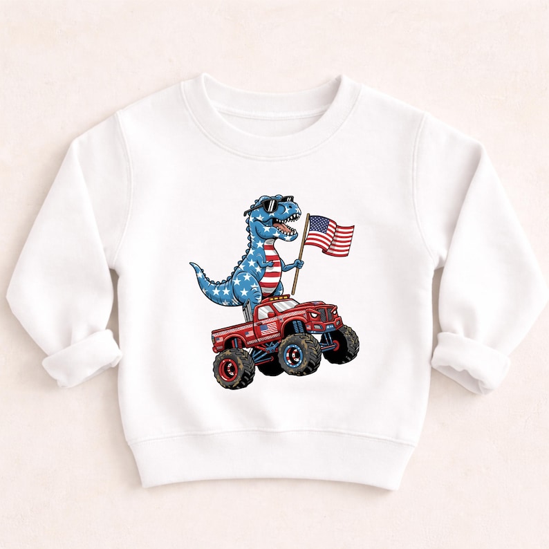A white long-sleeved shirt with a graphic design depicting a dinosaur riding a red monster truck while holding an American flag.