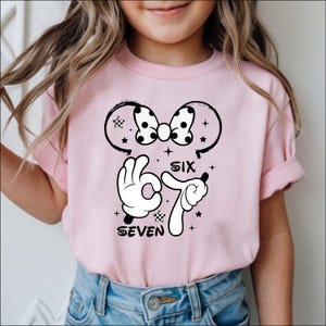 A young girl wearing a pink t-shirt with a cartoon character and text printed on it.
