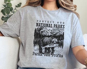 May include: Gray t-shirt with a black and white illustration of a bear holding a fish. The text "Protect Our National Parks Defund Doge Parks, Not Profits" is printed on the shirt.