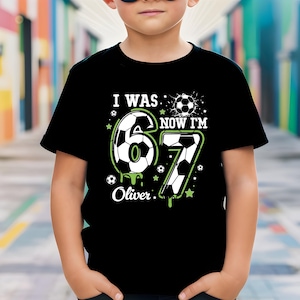 May include: Black t-shirt with a soccer-themed design for a 6-year-old's birthday. The shirt features the text "I WAS 6, NOW I'M 7" with the number 6 and 7 designed as soccer balls, and the name "Oliver".