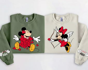 May include: Two sweatshirts, one olive green and one cream, featuring Minnie Mouse designs. One shows Minnie surrounded by heart shapes, the other as Cupid with a bow and arrow. Sleeves have a hand-drawn graphic and the letters "A&H".
