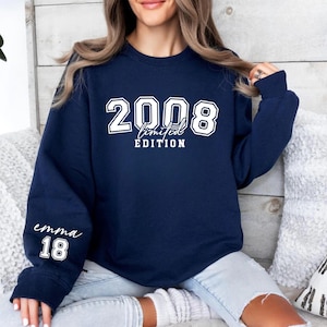 May include: Navy blue sweatshirt with the text "2008 Limited Edition" in white on the front. The sleeve features the name "emma" and the number "18" in white. The sweatshirt is a casual style.
