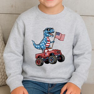 A child wearing a gray sweatshirt with a cartoon image of a dinosaur riding a red truck with American flags on the hood.