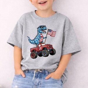 A young boy wearing a gray t-shirt with a graphic of a dinosaur riding a red truck and holding an American flag.
