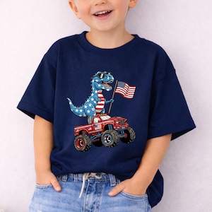 A young boy wearing a navy blue t-shirt with a graphic of a dinosaur riding a red monster truck and holding an American flag.