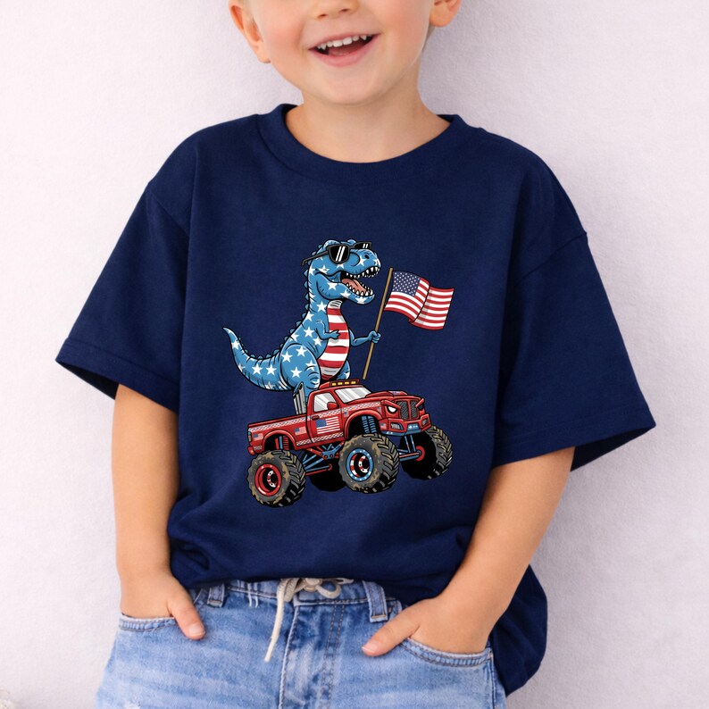 A young boy wearing a navy blue t-shirt with a graphic of a dinosaur riding a red monster truck and holding an American flag.