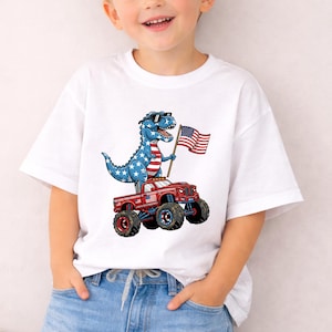 A young boy wearing a white t-shirt with a graphic of a dinosaur riding a red monster truck and holding an American flag.