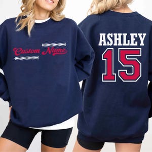 May include: Navy blue sweatshirt with the text "Custom Name" in red script and baseball stitching. The back of the sweatshirt has the name "Ashley" in white block letters above the number "15" in red with a white outline.