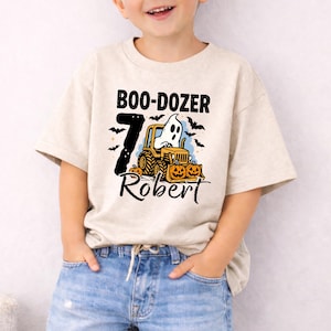 May include: A beige short-sleeve t-shirt featuring a graphic of a ghost driving a yellow tractor with pumpkins. The text reads BOO-DOZER, the number 7, and the name Robert. Black bats are scattered around the design on the front of the shirt.