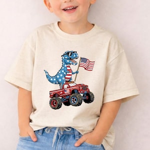 A young boy wearing a t-shirt with a cartoon dinosaur riding a red monster truck and holding an American flag.