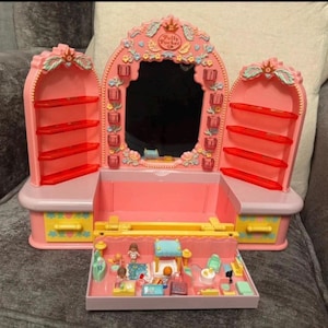 May include: A pink Polly Pocket playset featuring a mirror, shelves, and a miniature interior scene. The playset has a princess theme with floral and crown details. The interior scene includes miniature furniture and figures.