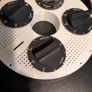 May include: A white circular appliance with four black control knobs labeled "OFF", "LOW", "MED", and "HI". The appliance has a series of small holes and a central opening. The knobs are set to various positions.