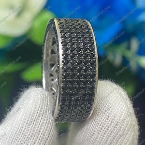 Puede incluir: Un anillo plateado con una banda cubierta de pequeñas piedras negras. El anillo está sostenido por una mano enguantada, con las piedras preciosas captando la luz. El diseño del anillo es una fila continua de piedras negras.