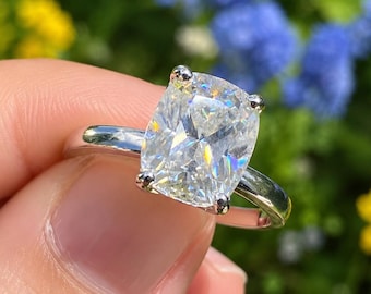 May include: A silver-toned ring featuring a large, clear, cushion-cut gemstone held in place by four prongs. The band is smooth and polished, showcasing a reflective metallic finish that contrasts with the faceted, brilliant-cut center stone.