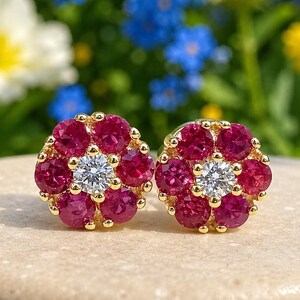 May include: A pair of flower-shaped stud earrings. Each earring features a central, round, clear gemstone surrounded by a cluster of deep red gemstones. The earrings are set in gold and rest on a light-colored surface.