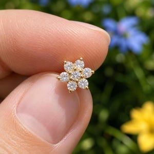 Könnte beinhalten: Ein goldener Ohrstecker in Blumenform mit sieben kleinen, klaren, rund geschliffenen Steinen in einer Cluster-Anordnung. Der mittlere Stein ist von sechs identischen Steinen umgeben, die alle in einer polierten Goldfassung mit Krappen sitzen.