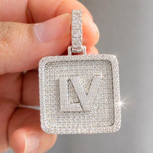 Può includere: Un ciondolo quadrato color argento con le lettere LV al centro, interamente incastonato con piccole pietre trasparenti. Il ciondolo presenta un bordo spesso tempestato di pietre e un gancio rettangolare sulla parte superiore.