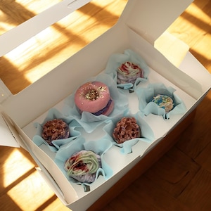 May include: A white cardboard box with a clear window containing five assorted bath bombs in light blue paper liners. The collection includes a pink donut-shaped bath bomb with gold glitter and four textured, multi-colored round bath bombs.