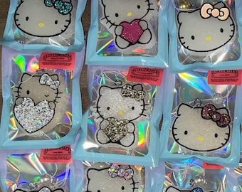 May include: Ten clear plastic bags with white Hello Kitty charms inside. Each charm is a different color and has a different design. The charms are all sparkly and have a heart-shaped design. The bags are all labeled with the text "Freshnie Scents".