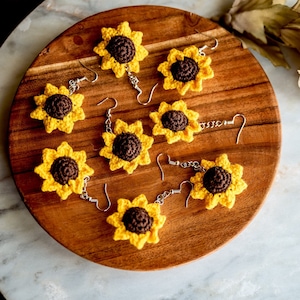 May include: Six pairs of crocheted sunflower earrings on a wooden plate. The earrings are yellow and brown with silver hooks.