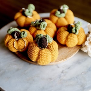 May include: Six crocheted pumpkin keychains in orange with green stems and brown tops. The keychains are arranged on a wooden plate on a marble surface.