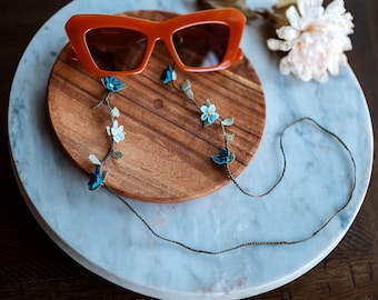 May include: Orange cat-eye sunglasses with a floral beaded chain. The chain has blue and white flowers and green leaves.