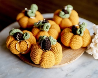 May include: Six crocheted pumpkin keychains in orange with green stems and brown tops. The keychains are arranged on a wooden plate on a marble surface.