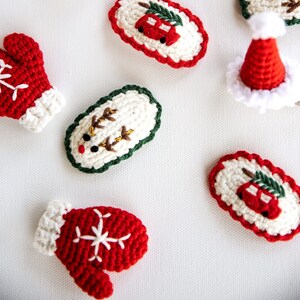 Puede incluir: Un conjunto de cinco adornos navideños de ganchillo. Los adornos son blancos con detalles rojos y verdes. Un adorno tiene un reno, otro tiene un coche con un árbol de Navidad en la parte superior, y dos son guantes. El quinto adorno es un gorro de Papá Noel rojo.