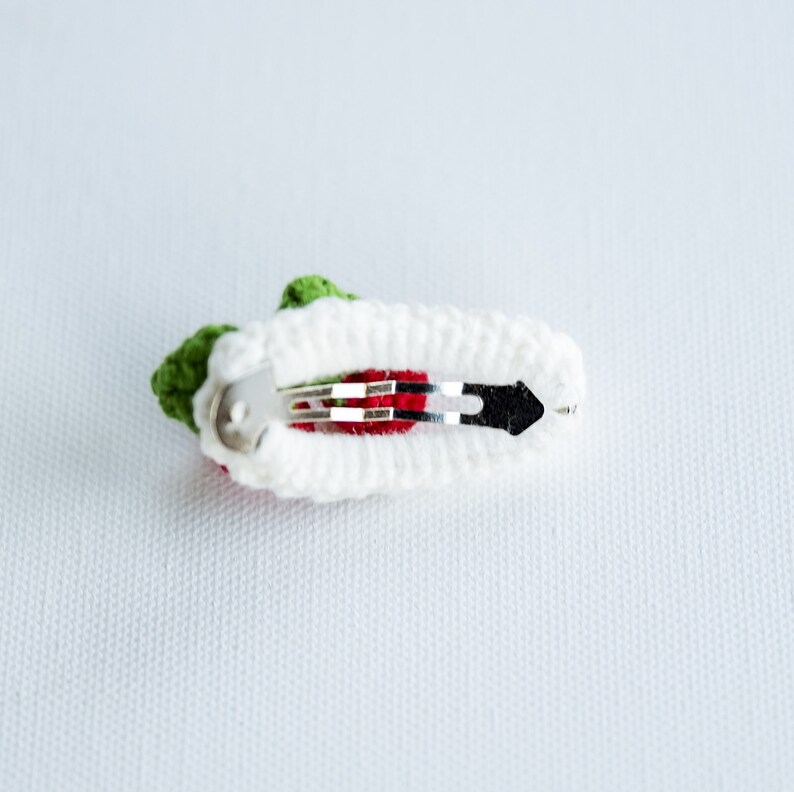 Puede incluir: Una pinza para el pelo de crochet blanco con un dise&ntilde;o de flor roja y verde. La pinza tiene un cierre de metal plateado.