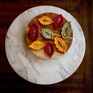 May include: A set of eight crocheted leaf-shaped hair clips in shades of yellow, red, and green. The clips are arranged on a wooden tray with a marble surface.