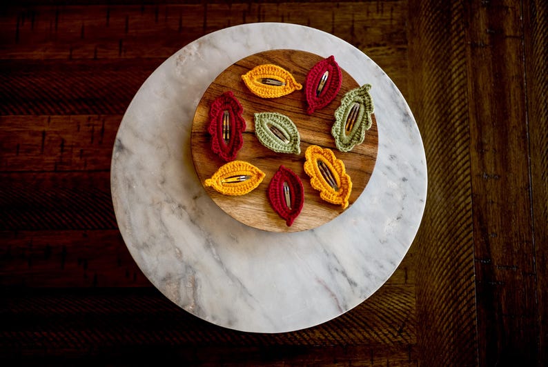 May include: A set of eight crocheted leaf-shaped hair clips in shades of yellow, red, and green. The clips are arranged on a wooden tray with a marble surface.