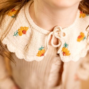 May include: Cream-colored knitted collar with embroidered yellow and orange flowers and green leaves. The collar has a scalloped edge and a tie closure at the front. The garment is likely for a child.