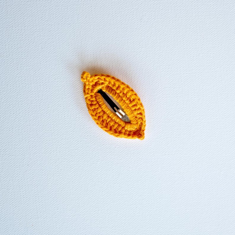 May include: A small, crocheted hair clip in the shape of a leaf. The clip is a bright orange color and has a metal clasp.