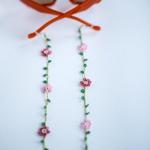 Pu&ograve; includere: Un paio di occhiali da sole arancioni con una catena floreale di perline. La catena ha fiori rosa e bianchi con foglie verdi.