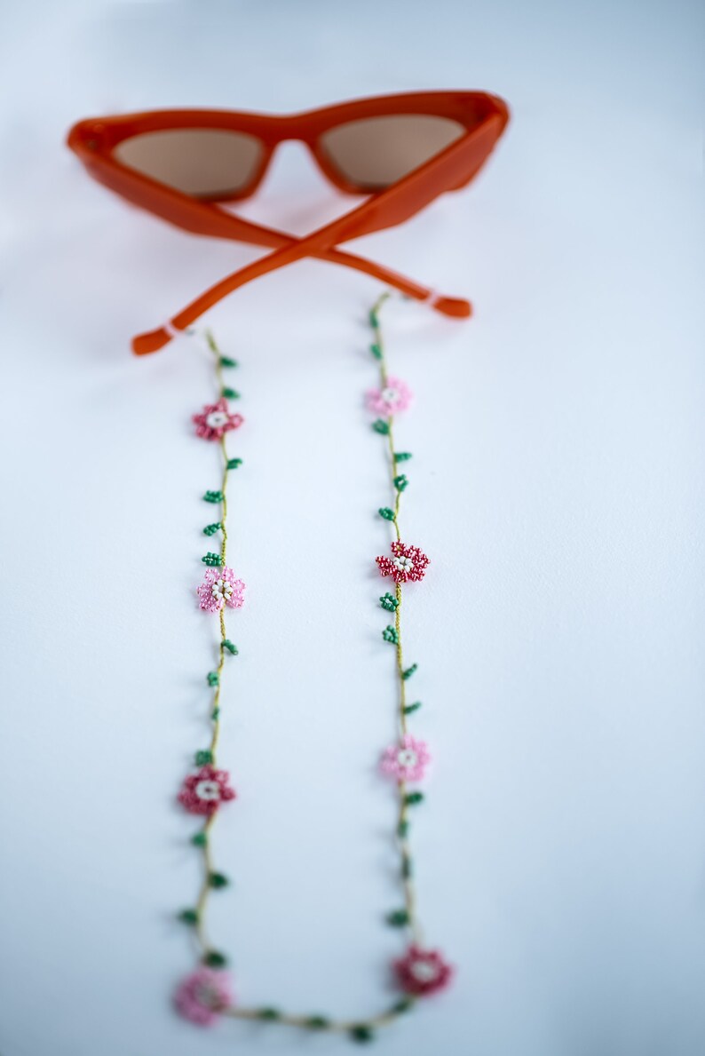 Pu&ograve; includere: Un paio di occhiali da sole arancioni con una catena floreale di perline. La catena ha fiori rosa e bianchi con foglie verdi.